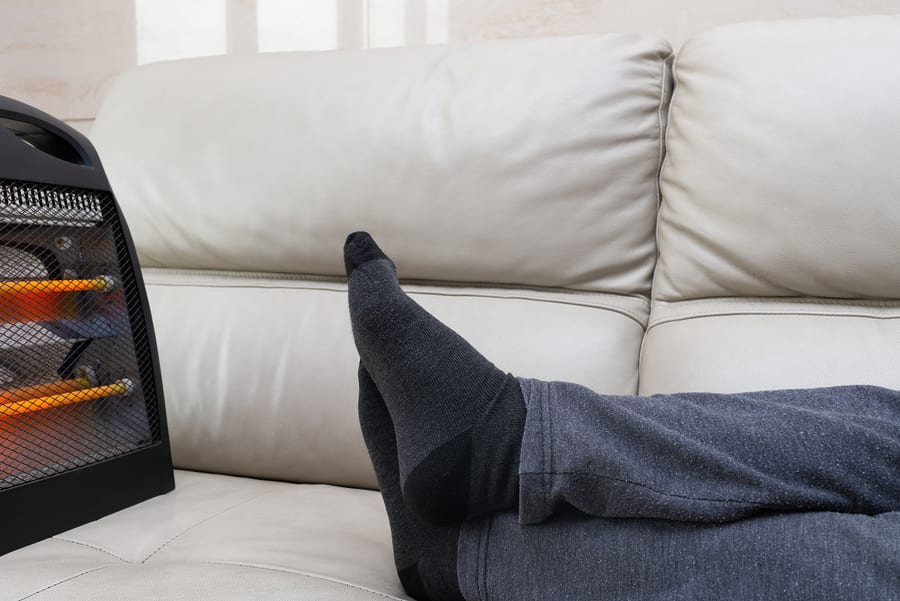 Man Lying On Sofa With An Electric Heater Near His Feet